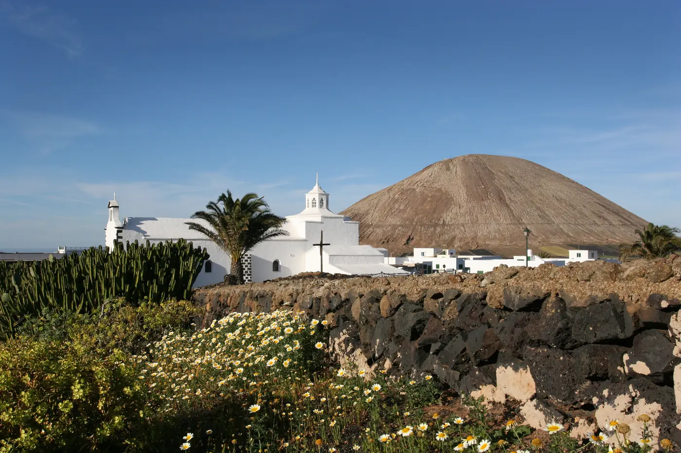 The white church of Los Dolores in Mancha Blanca behind a lava-stone wall and daisies, with a volcanic cone beyond.