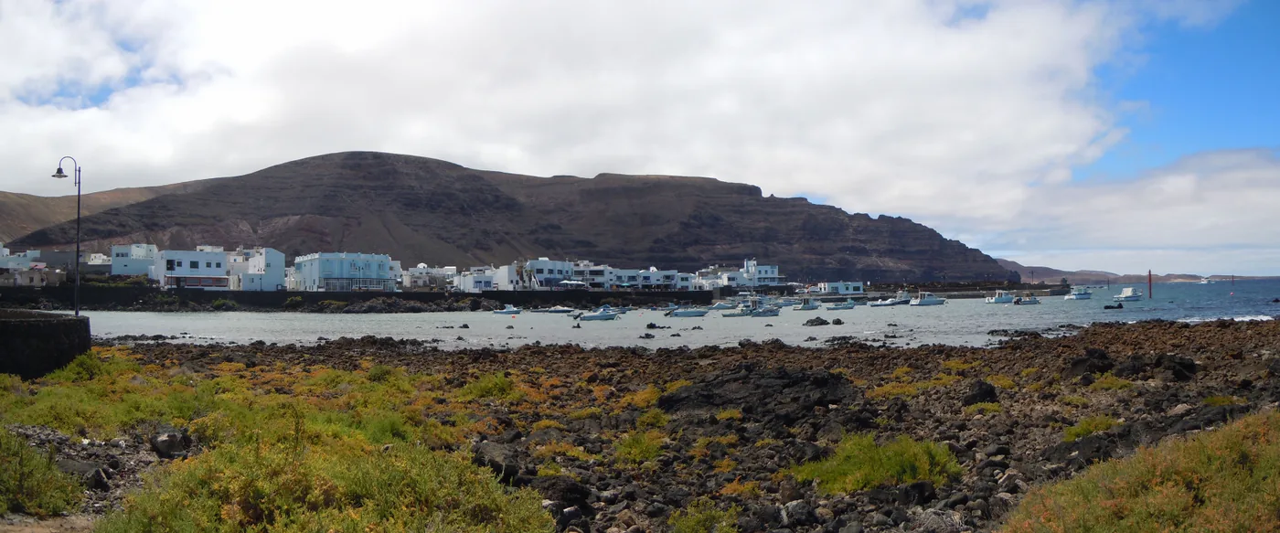 Órzola's white houses and harbour seen across a low lava shore, below dark hills.