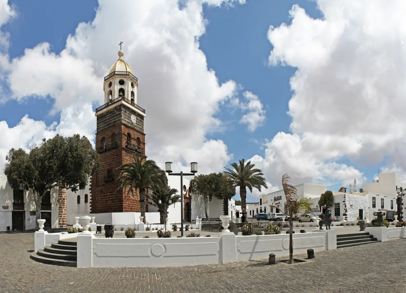 The brick bell tower of the church of Nuestra Señora de Guadalupe beside the white houses and palms of Plaza de la Constitución in Villa de Teguise, under a blue sky.