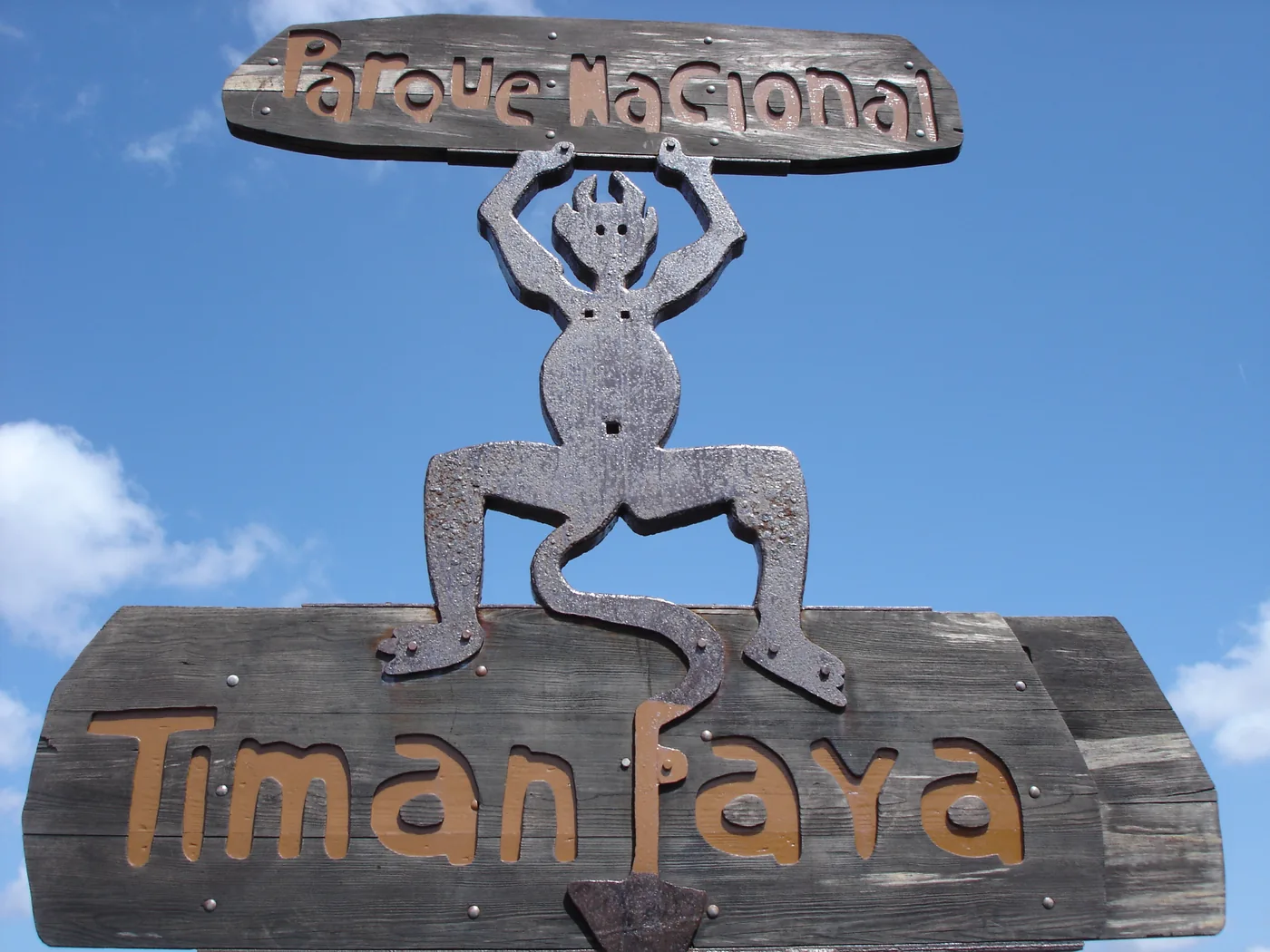 Timanfaya's metal devil emblem on a wooden sign reading Parque Nacional Timanfaya, against a blue sky.
